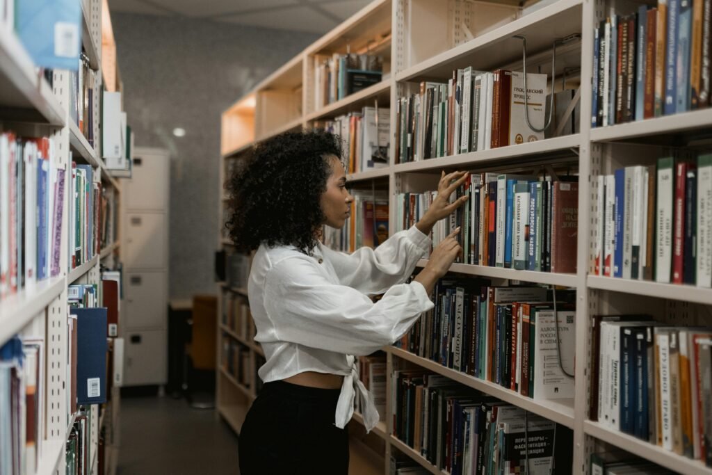 A young woman with curly hair searches for books on a library bookshelf.
